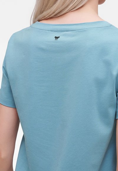 Personne blonde portant une chemise bleu clair à manches courtes avec un petit logo papillon noir et le texte "WN" sur le haut du dos.