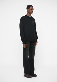 Pulover negru tricotat cu guler rotund și detalii subtile cu logo; asortat cu pantaloni din denim închis și încălțăminte neagră. Design minimalist.