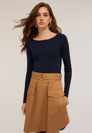 Vrouw met lang golvend haar, die gouden oorbellen draagt, een marineblauwe geribde lange mouw top en een beige rok met grote voorzakken en een riem.