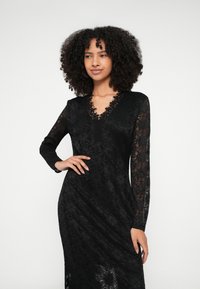 Crna čipkana haljina s dugim rukavima, V-izrezom, složenim cvjetnim uzorcima, uskim krojem i detaljima s čipkanim obrubom. Mat završna obrada s prozirnim naglascima.