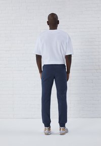 Vit t-shirt med kort ärm och marinblåa sweatpants med en bakficka, i kombination med grå och gula sportskor mot en vit tegelbakgrund.