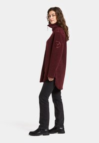 Bordeaux-fleece pusero, jossa on korkea kaulus ja epäsymmetrinen helma. Tekstuuroitu pinta ja logo-patch vasemmassa hihassa. Mustat farkut ja saappaat.