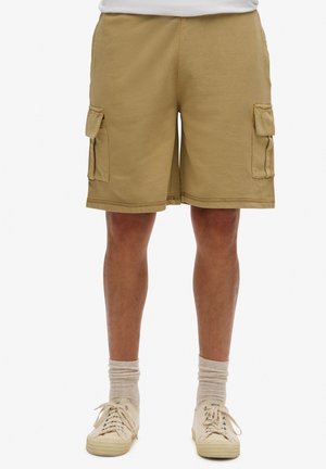 Shorts cargo arrivés au genou en tissu kaki, dotés de deux poches latérales. Portés avec des baskets beiges et des chaussettes mi-mollet grises claires.