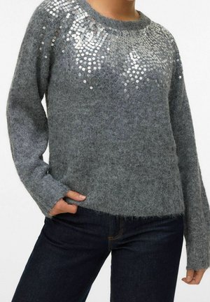 Grå, blød sweater med sølvfarvede pailletter på bryst og skuldre, båret med mørkeblå jeans, hånd i lommen.