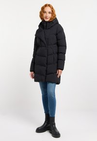 Abrigo de plumas negro con capucha, que cuenta con cierre de cremallera y detalles de botones. Lleva puesto sobre vaqueros azules y botas de tobillo, destacando una silueta moderna.