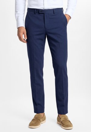 Homme portant un pantalon bleu marine slim fit, une chemise blanche, des mocassins en daim beige, debout avec une main dans la poche sur fond blanc.