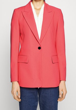 Personne portant un blazer corail vif à un bouton sur un haut blanc zippé et un jean bleu foncé, debout devant un fond uni.