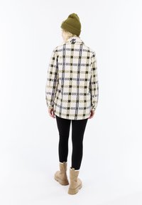 Persona con un gorro verde, chaqueta blanca y verde con estampado, leggings negros y botas beige, de pie de espaldas a la cámara.