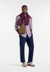 Borsa crossbody in tessuto beige, camicia button-up azzurro chiaro, pantaloni blu navy, maglione bordeaux avvolto intorno alle spalle, sneakers grigie con dettagli.