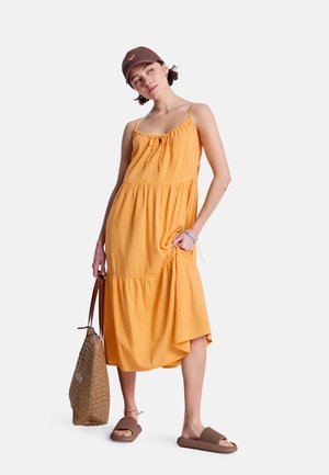 Żółta bawełniana sukienka warstwowa na cienkich ramiączkach, z marszczonym dekoltem i boczną kieszenią. Do tego brązowe sandały i dopasowana czapka. Pleciona torba na ramię.