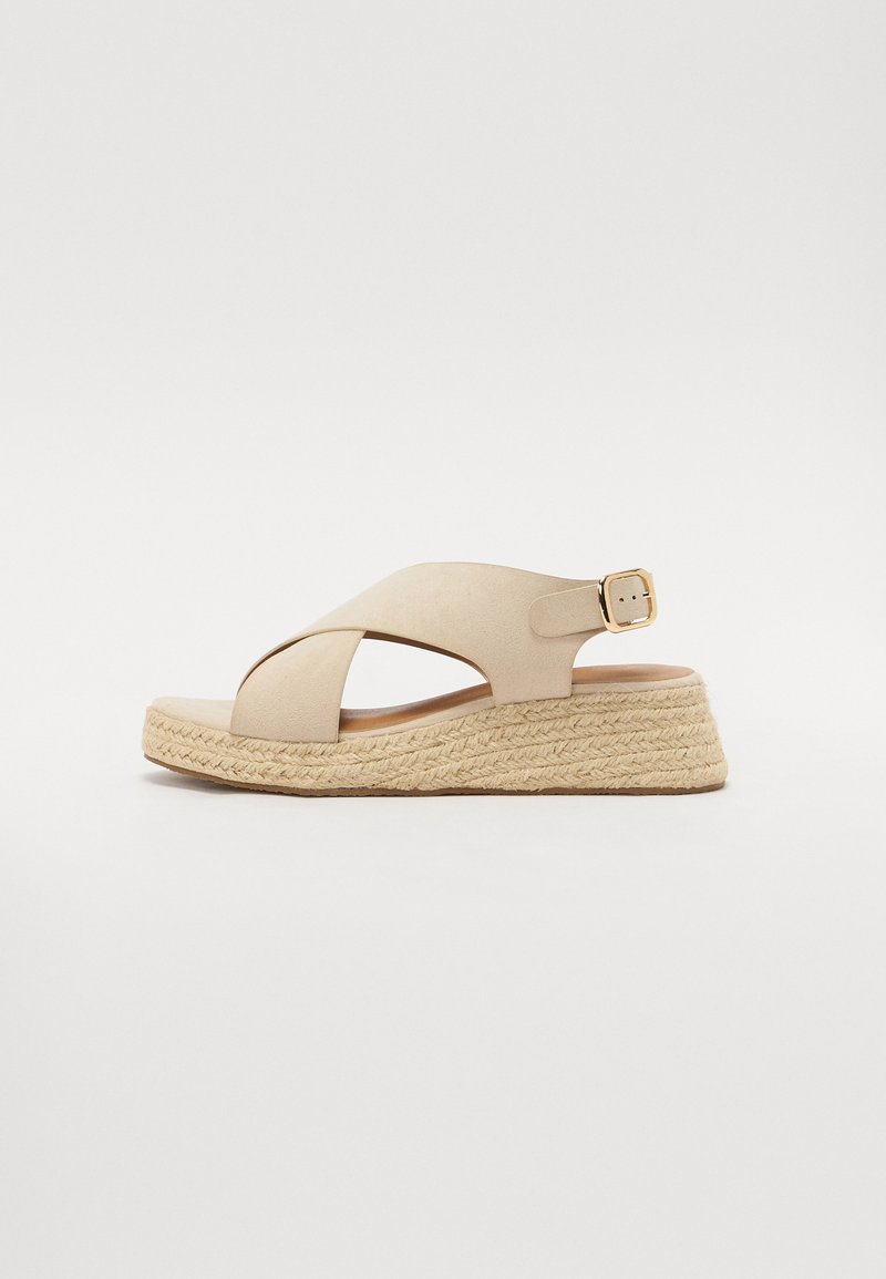 Beige espadrille sandaalit, joissa on ristiin menevät hihnat, korokepohja juutista ja kultainen solkiyksityiskohta. Pehmeä pinta ja yksinkertainen muotoilu.