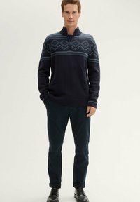 Granatowy sweter z zamkiem na ćwierć, z geometrycznym wzorem w jaśniejszym odcieniu niebieskiego na klatce piersiowej, w zestawie z ciemnymi spodniami i butami.