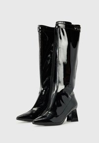 Bottes noires en cuir verni, montantes jusqu'au genou, avec des bouts pointus et des talons blocs, dotées d'une fermeture éclair avant et d'accents décoratifs en métal.