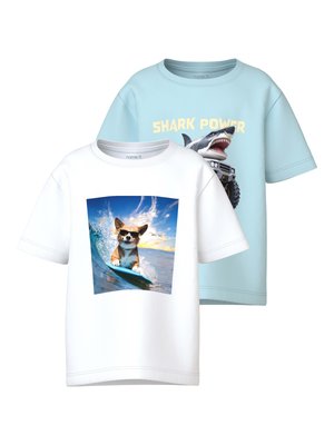 Λευκό μπλουζάκι με σκύλο που φοράει γυαλιά ηλίου και σερφάρει σε κύμα· ανοιχτό μπλε μπλουζάκι με καρχαρία και τον κείμενο "SHARK POWER" πάνω από ένα τερατότροχο φορτηγό.