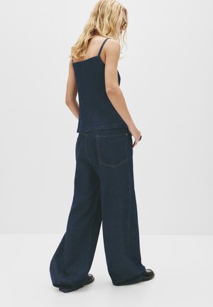 Dark blue denim top with thin straps and a structured shape, paired with wide-legged dark blue denim pants featuring back pockets and orange stitching.