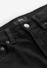 Jeans de mezclilla negra con una cintura cosida, cierre de botón plateado y etiqueta de tela marcada "NEXT". Tejido texturizado con costuras visibles.