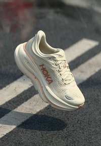 Λευκό και ροδακινί παπούτσι τρεξίματος Hoka στον αέρα πάνω από άσφαλτο με λευκές γραμμές δρόμου και μικρά συντρίμμια γύρω από τη σόλα.