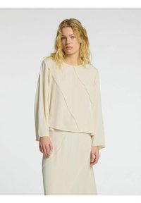 Cremefarvet langærmet top med løs pasform, der har rå kanter og skrå sømme. Glat tekstur, lagdelt design og minimalistisk stil.
