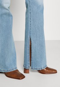 Helleblaue Schlagjeans mit rückwärtigen Knöchelschlitzen, getragen über braunen Stiefeletten im Krokodilmuster mit Absatz.