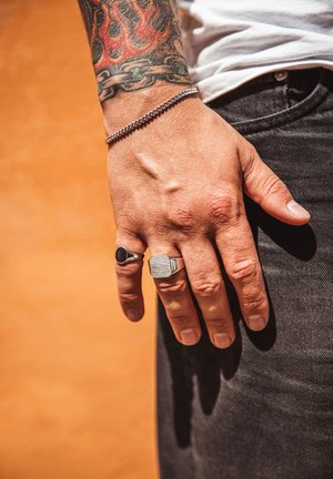 Sølvstænger med matte og glatte overflader på fingrene, et sølvkædearmbånd, og mørk denim, med livlige tatoveringsdetaljer synlige.