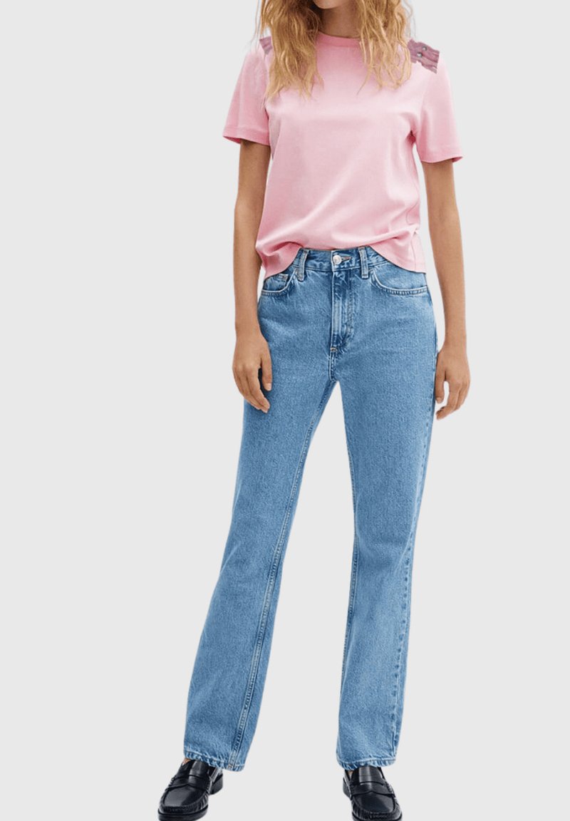 Camiseta de color rosa claro con mangas cortas y detalles texturizados en los hombros, combinada con jeans azul denim de pierna recta y mocasines negros.