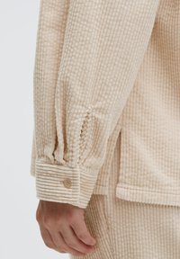 Cremfarbene Corduroy-Hemd mit gerippter Textur, Raffung an den Ärmeln und Knopfmanschette. Bietet eine lockere Passform und einen Seitenschlitz für mehr Bewegungsfreiheit.