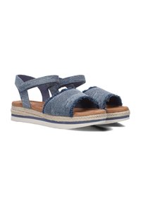 Denimblauwe sandalen met bandjes en rafelige randen. De zool heeft een geweven jute accent en een witte rubberen basis met een blauwe streep. Lichtgewicht ontwerp.