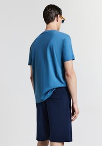 T-shirt blu a maniche corte in un tessuto morbido, abbinato a pantaloni corti navy con una tasca posteriore, mostrato da dietro. Occhiali da sole inclusi.