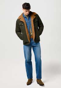 Chaqueta verde con cuello de piel de oveja marrón, combinada con una camisa de mezclilla azul y jeans azul claro. Botas de cuero marrón completan el conjunto.