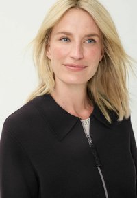 Schwarzer Cardigan mit Reißverschluss, spitzem Kragen, strukturiertem Stoff und einem silbernen Reißverschluss. Weiches, minimalistisches Design mit subtiler Schattenspiel.