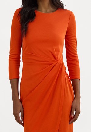Femme portant une robe orange vif ajustée avec des manches trois-quarts et un détail de nœud sur le côté à la taille.