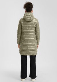 Olivovo zelená prešívaná bunda s kapucňou, horizontálnym švom, strednej dĺžky, lesklý povrch. Skombinovaná s čiernymi rozšírenými nohavicami a topánkami.