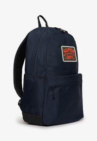 Mochila navy con una tela texturizada, que presenta un logo en parches en rojo y blanco, un bolsillo frontal y un acento negro en la base.
