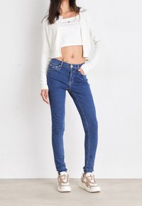 Vaqueros skinny azules de cintura alta, combinados con un cárdigan blanco corto y una camiseta sin mangas. Lleva zapatillas chunky. Tejido suave, ajuste casual.