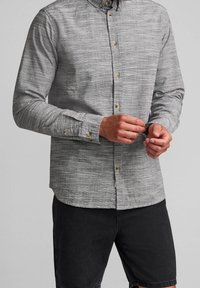 Camicia button-up grigia con texture e maniche lunghe, caratterizzata da bottoni in ottone, indossata in modo casual su pantaloni corti neri.