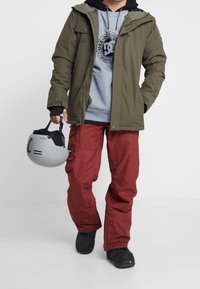 Outfit da snowboard con giacca verde oliva, felpa grigia, pantaloni rossi e stivali neri. Tiene un casco grigio con accenti neri.