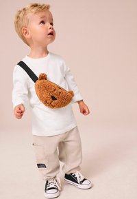 Vit långärmad skjorta, beige byxor, svarta sneakers. Brun plysch axelväska formad som en björn, med en texturerad yta och ansiktsdetaljer.