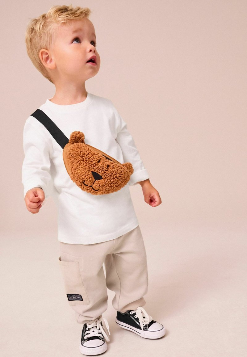 Vit långärmad skjorta, beige byxor, svarta sneakers. Brun plysch axelväska formad som en björn, med en texturerad yta och ansiktsdetaljer.