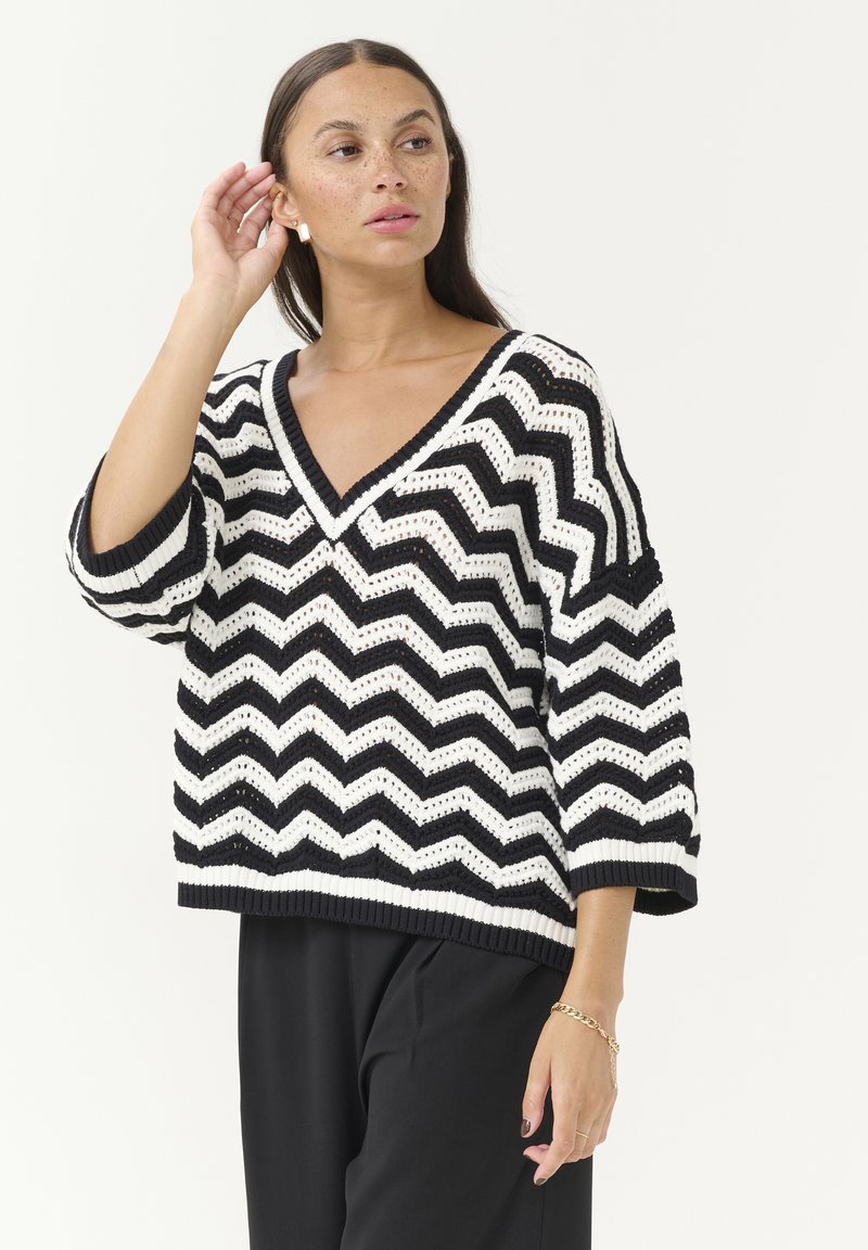 Pull en tricot à motif chevron noir et blanc avec un col en V, coupe ample, manches trois-quarts et ourlet côtelé. Tissu texturé.