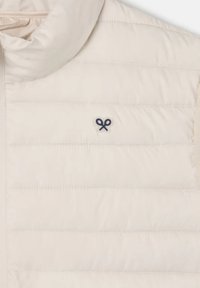 Chaleco acolchado beige con cuello alto y pequeño logotipo azul marino; textura suave, costuras horizontales, diseño ligero.