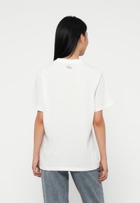 Mujer con cabello largo y negro de espaldas, vistiendo una camiseta blanca sencilla con un pequeño texto rojo cerca del escote y vaqueros azules.