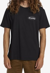 Mężczyzna w czarnej koszulce z krótkim rękawem z małym białym logo "Billabong" na lewej piersi i brązowych spodniach, stojący na tle białym.