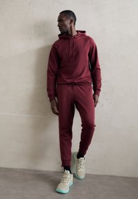 Bordowa bluza z kapturem i dresy wykonane z miękkiego materiału, związywane sznurkiem i zakończone ściągaczami, w połączeniu z beżowymi sneakerami i ciemnymi skarpetami.