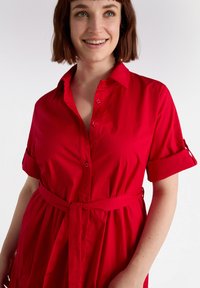 Red shirt dress with a collar, short rolled sleeves, button front, and a tied waist belt. Smooth fabric with a relaxed fit.