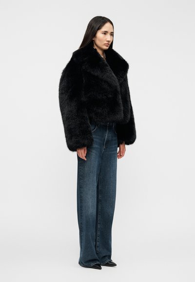 Μαύρο μπουφάν από ψεύτικη γούνα με πλατύ γιακά, σε συνδυασμό με φαρδιά μπλε τζιν. Το σύνολο ολοκληρώνεται με μυτερά μαύρα παπούτσια.