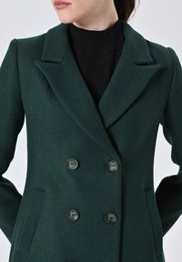 Cappotto doppio petto verde scuro realizzato in tessuto testurizzato, con colletto a punta e due grandi bottoni sul davanti.