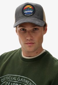 Graue Cordhut mit beigefarbenem Netzrücken, mit einem runden Aufnäher, der Berge und einen Sonnenuntergang in lebhaften Farben zeigt. Darunter ein grüner Sweatshirt.