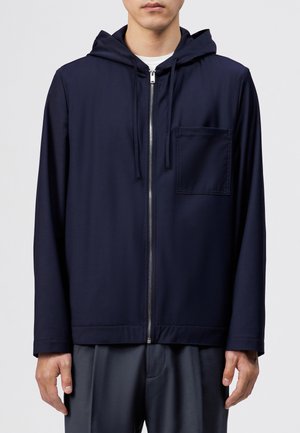 Mand iført en marineblå lynlåshættejakke med brystlomme og snoretræk, kombineret med mørke bukser og hvid inderskjorte.