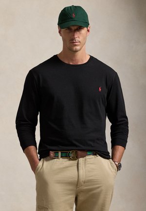 Uomo che indossa una maglietta nera a maniche lunghe, pantaloni beige con cintura a righe verdi, berretto verde scuro e orologio da polso, in piedi con le mani in tasca.