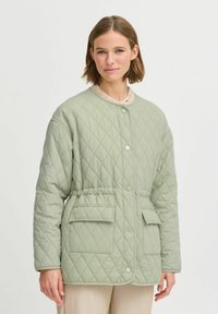 Chaqueta acolchada en verde claro, con cuello redondo, cierre de botones en el frente, dos bolsillos delanteros y una cintura elástica para un ajuste ceñido.